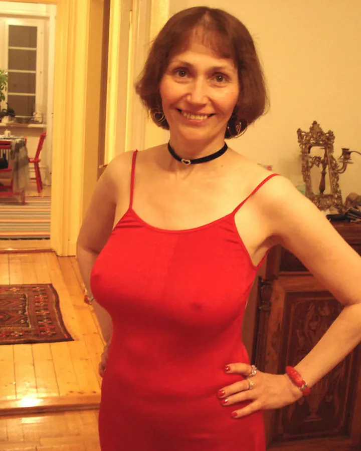 Femme aux cheveux bruns courts, souriante, portant une robe rouge ajustée, debout dans un intérieur avec un meuble en bois sculpté et des bougies rouges.