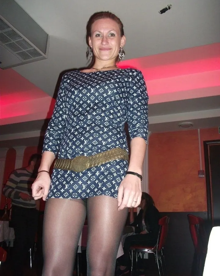 Femme aux cheveux châtains, souriante, en robe à motif monogramme bleu foncé, avec ceinture dorée et collants noirs dans un lieu intérieur éclairé en rose.