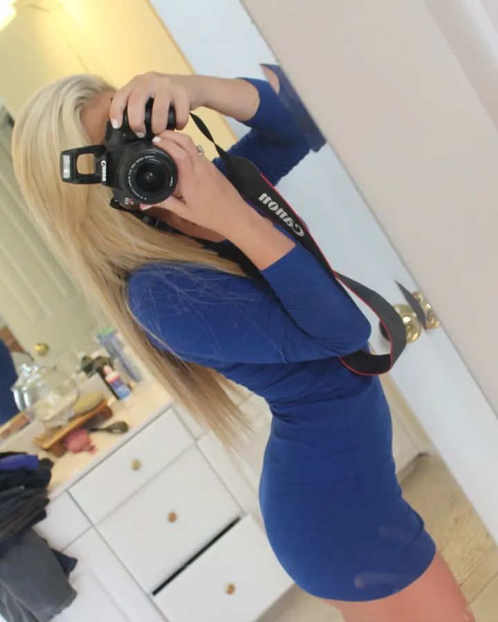 Femme aux cheveux blonds platine portant un haut noir et des collants noirs, assise sur un canapé